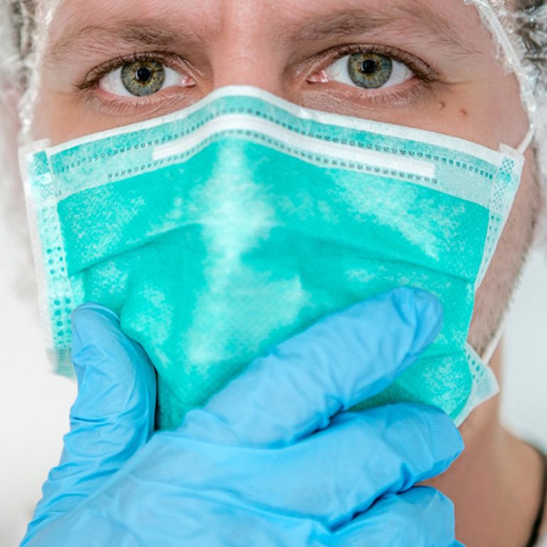 männliches Gesicht mit grünen Augen, der Mannn trägt eine OP-Haube OP Masken und blauen Klinik Schutzhandschuhen